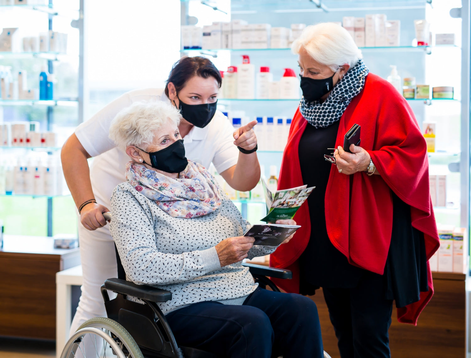 Eine ältere Dame im Rollstuhl wird von einer Pflegekraft begleitet und bespricht Medikamentenbroschüren mit einer Apothekerin in einer Apotheke.